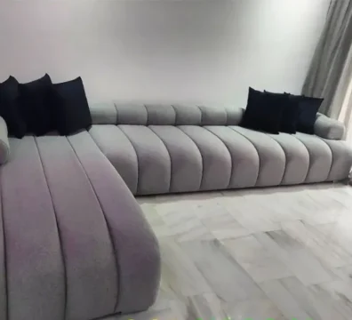 Canapé d'angle au design unique avec un capitonnage vertical tubulaire sur toute la structure, en tissu gris clair, pour un salon contemporain. Ameublement Hicham.