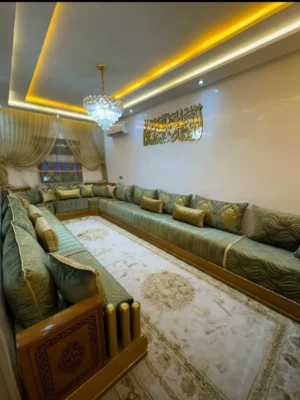 Grand salon marocain panoramique en velours vert sauge, avec des coussins dorés et une structure en bois avec finition dorée. Ameublement Hicham Marrakech.