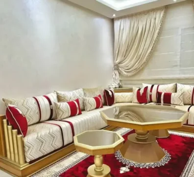Salon marocain moderne en L avec une structure dorée, un tissu blanc à motifs géométriques et des coussins décoratifs rouges. Ameublement Hicham Marrakech.