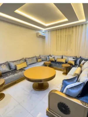 Salon marocain moderne en U avec sedari en tissu bleu et détails dorés, tables d'appoint et structure en bois doré. Ameublement Hicham Marrakech.