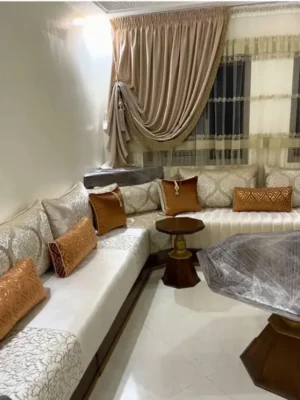 Salon marocain en L avec un élégant tissu damassé blanc cassé, rehaussé de coussins décoratifs couleur terracotta. Ameublement Hicham Marrakech.
