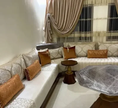Salon marocain en L avec un élégant tissu damassé blanc cassé, rehaussé de coussins décoratifs couleur terracotta. Ameublement Hicham Marrakech.