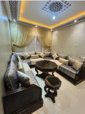 Salon marocain traditionnel en U avec un tissu à motifs sombres et des tables en bois foncé, sous un plafond décoré de gebs. Ameublement Hicham Marrakech.