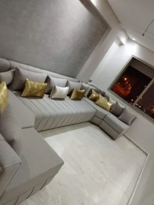 Salon moderne en U en tissu gris clair, avec une section à capitonnage vertical et des coussins dorés décoratifs. Ameublement Hicham Marrakech.